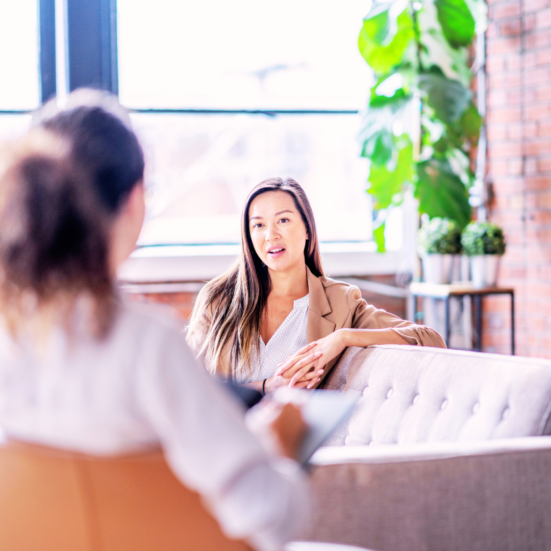 Two women sitting on a couch in a modern office setting with plants and large windows. Both are happy