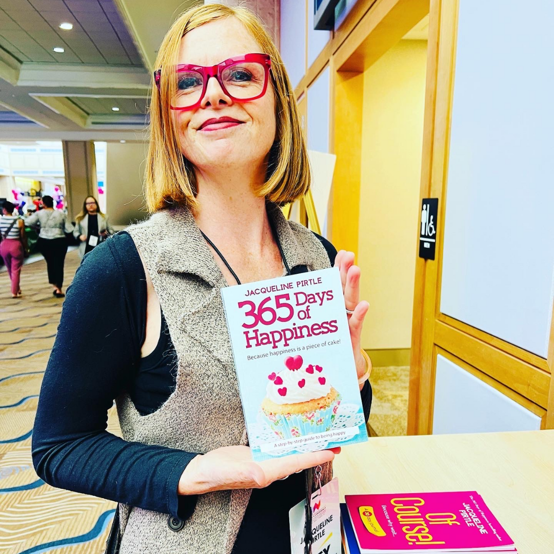 Author and healer Jacqueline Pirtle holding her bestselling book titled '365 Days of Happiness' in an indoor setting. She is smiling and happy.
